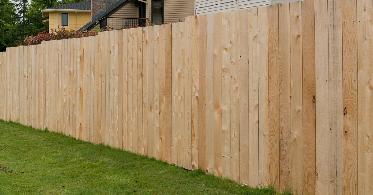 An unstained cedar wood picket fence in a grassy backyard. Behind the fence are multiple suburban homes in a line.