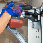 A repairman drills a screw into the frame of a residential garage door, wearing blue and orange work gloves.