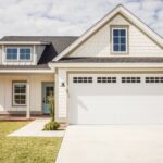 A suburban one-story house with white siding and a garage door featuring an empty driveway and a trimmed front lawn.