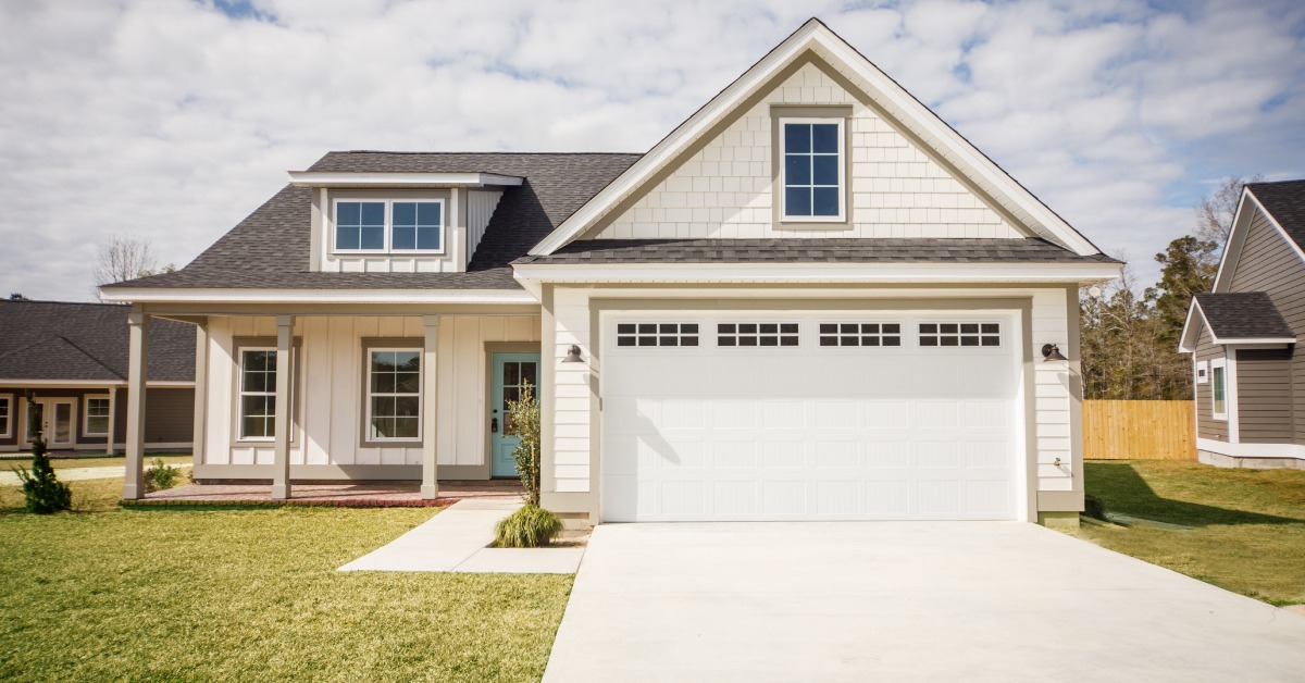 A suburban one-story house with white siding and a garage door featuring an empty driveway and a trimmed front lawn.