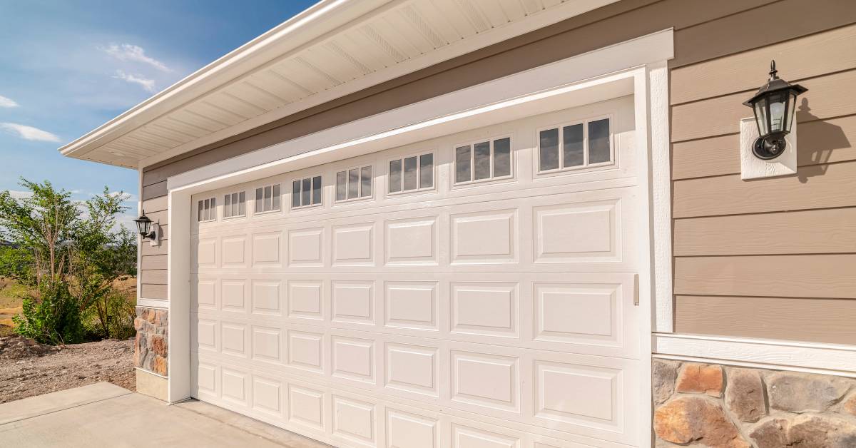 A light-colored garage door with windows on a home with beige siding and natural stone in sunny weather.