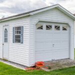 A white shed in the middle of a grassy backyard with an entry door, two windows, and a side overhead door.