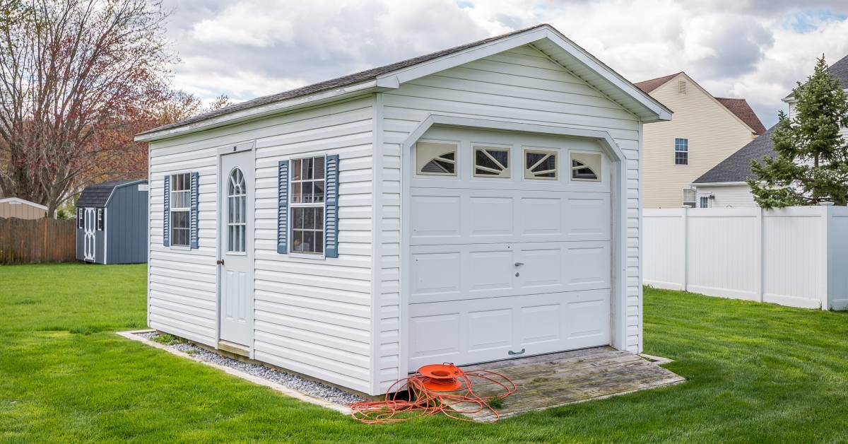 A white shed in the middle of a grassy backyard with an entry door, two windows, and a side overhead door.