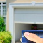 A person's hand holding a blue garage door opener remote to lift a garage door in the blurred background.