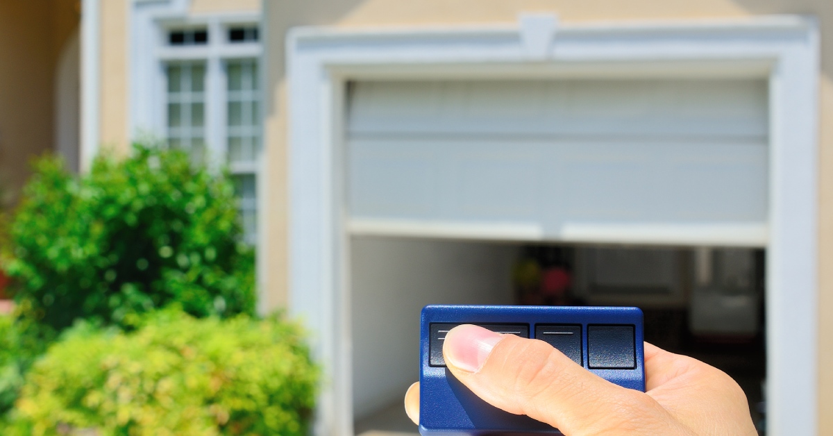 A person's hand holding a blue garage door opener remote to lift a garage door in the blurred background.