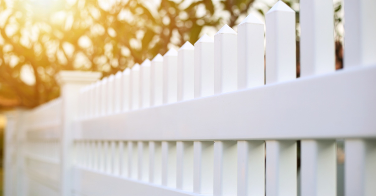 A close-up of a white picket residential vinyl fence with the sun shining through blurry tree branches.