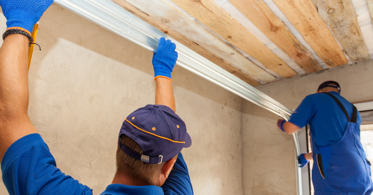 Two professional technicians wearing blue coveralls install a garage door track into an unfinished garage.