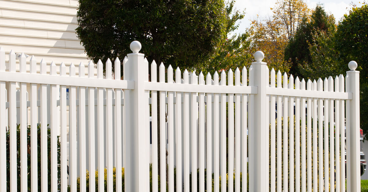 A vinyl white picket fence on a property with a cream-colored house. A sidewalk runs parallel to the fence leading to a driveway.