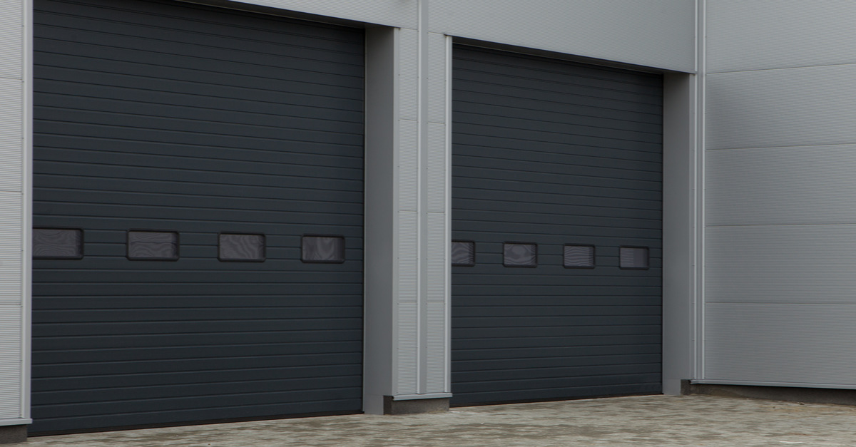 Two dark gray commercial roll-up garage doors are on a tall building. The doors feature one row of window panels each.