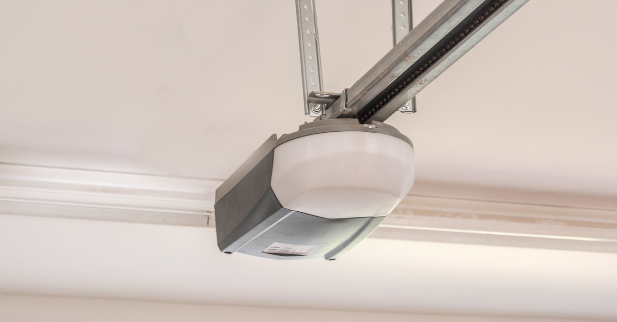 A gray garage door opener is mounted to a white ceiling, with a visible bracket and plastic cover on the unit's underside.