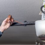 A bare hand uses a brown oil can on a black garage door opener chain hanging from a white textured ceiling.