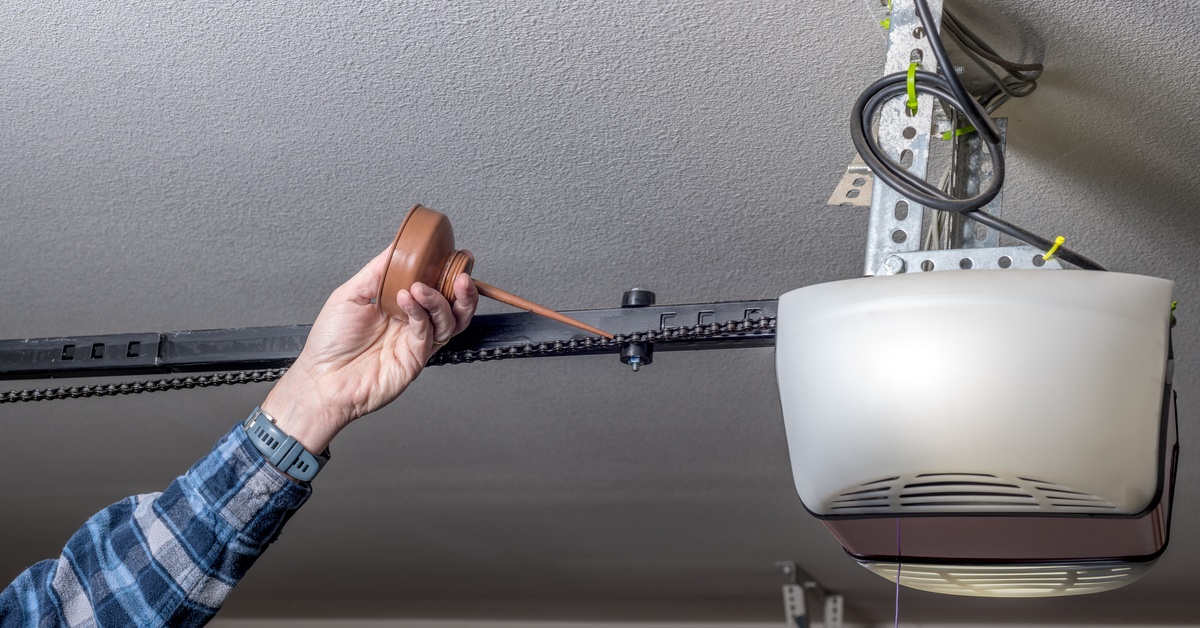 A bare hand uses a brown oil can on a black garage door opener chain hanging from a white textured ceiling.