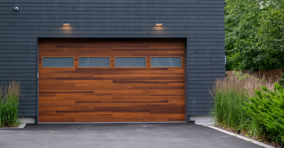 A faux wood garage door featuring horizontal frosted windows. Tall green plants border the door and concrete driveway.