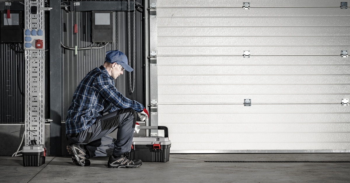 A man in goggles and gloves squats beside a white commercial garage door with a toolbox at his side.