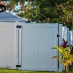 A solid white vinyl fence with black hinges stands on a green lawn beside a beige house with potted and planted greenery.