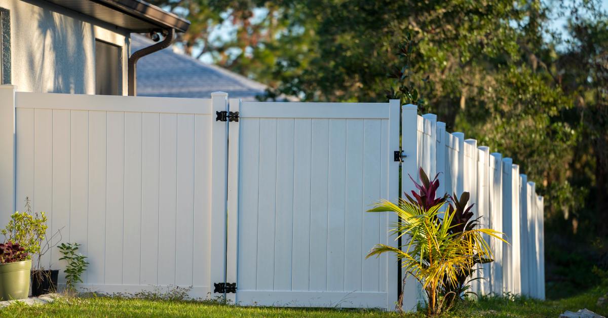 A solid white vinyl fence with black hinges stands on a green lawn beside a beige house with potted and planted greenery.