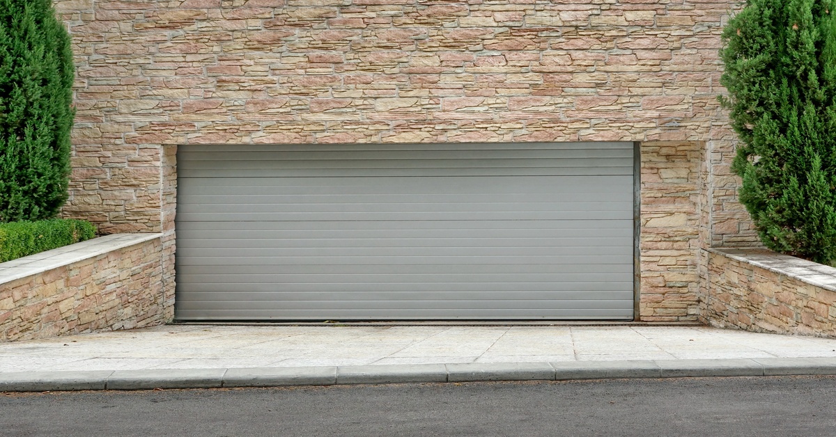 A gray roll-up commercial garage door sits at the base of a sloped driveway with stone retaining walls and greenery.