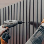 A worker wearing gloves uses a drill to install dark fence panels. Another worker holds some of the pieces in place.