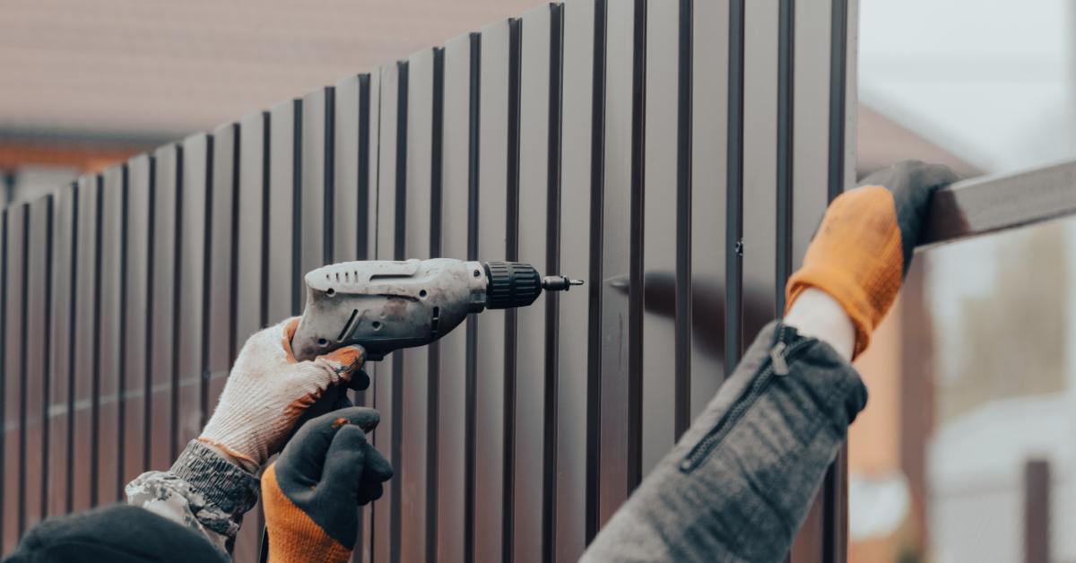 A worker wearing gloves uses a drill to install dark fence panels. Another worker holds some of the pieces in place.