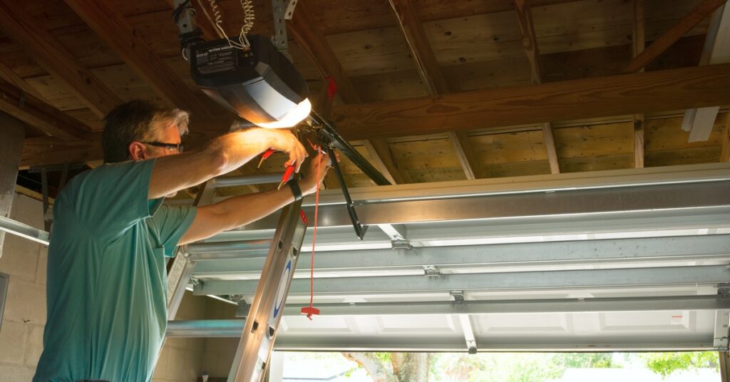 A man wearing glasses and a teal shirt stands on a ladder as he fixes the interior parts of a home's garage door.