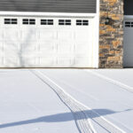 The front of two white garage doors on a home. The driveway is covered in snow, with tire tracks coming from the garage door.