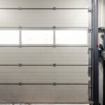 A man wearing a gray sweatshirt and workpants stands inside of a warehouse and opens a garage door.