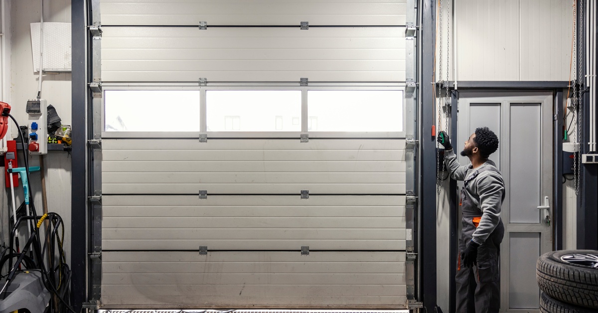 A man wearing a gray sweatshirt and workpants stands inside of a warehouse and opens a garage door.