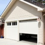 A cream-colored detached garage with a white garage door opens to show a dark interior and the bumper of a vehicle.