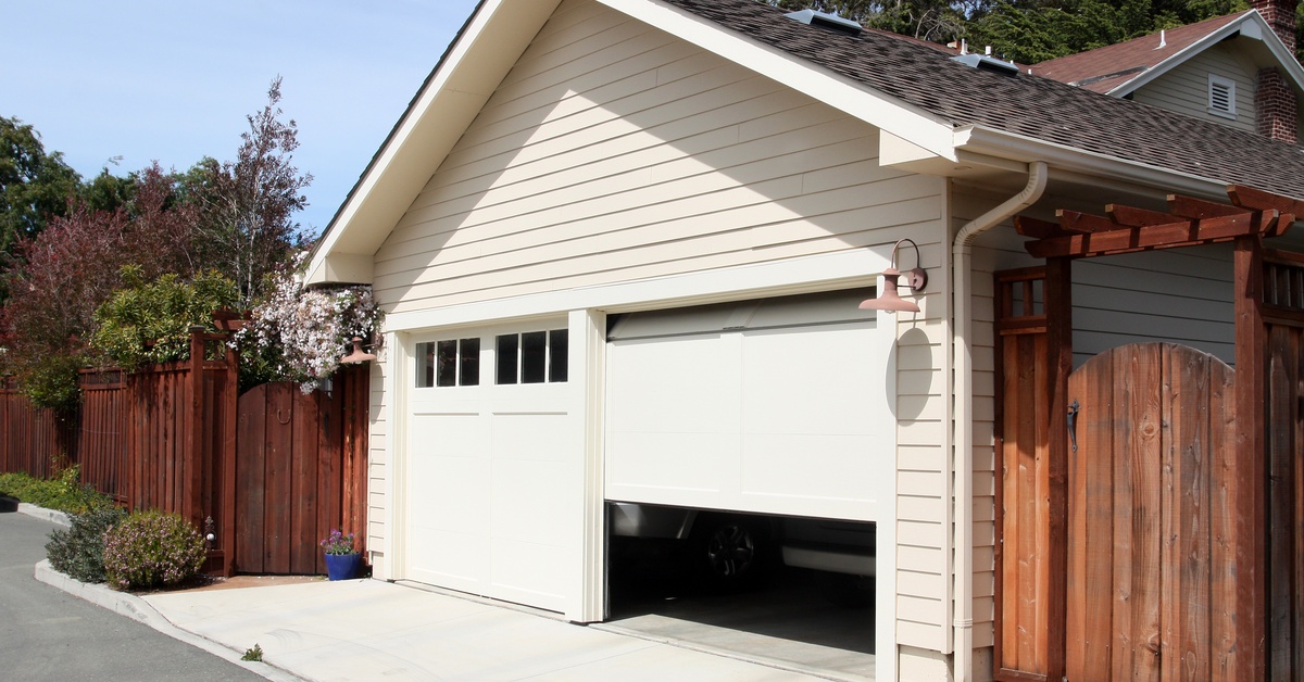 A cream-colored detached garage with a white garage door opens to show a dark interior and the bumper of a vehicle.