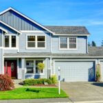 A two-story gray suburban home contains double garage doors, a red front door, green lawn, and white SUV in the driveway.