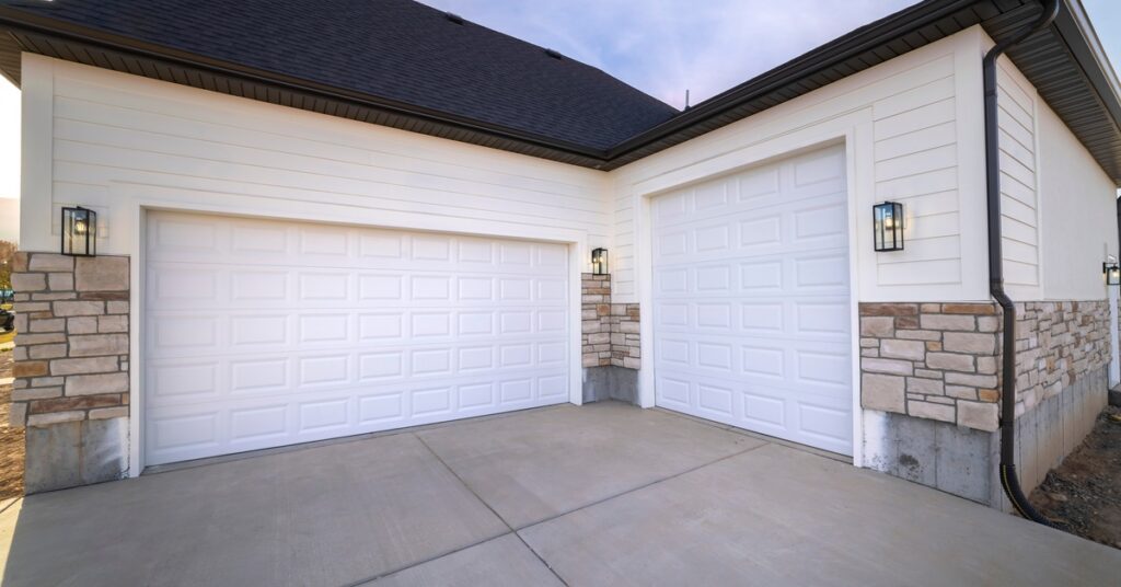 A beige home features two white paneled garage doors with stone accents above a clean concrete driveway