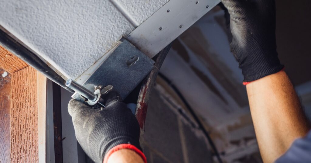 A technician wearing black gloves tightens a garage door track bracket with a wrench while standing inside the garage.