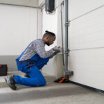 A garage door technician kneels beside a white sectional door, adjusting the track hardware with tools.