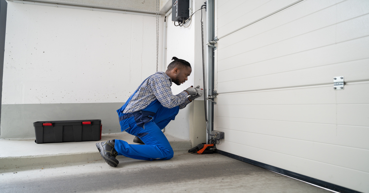 A garage door technician kneels beside a white sectional door, adjusting the track hardware with tools.