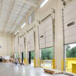 A warehouse contains partially-opened large industrial overhead doors with a forklift and pallets nearby.