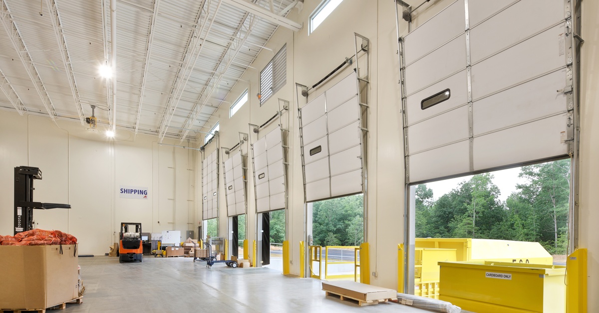 A warehouse contains partially-opened large industrial overhead doors with a forklift and pallets nearby.