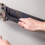 A hand holds a wrench against a coiled garage door spring attached to a metal track on a textured wall.
