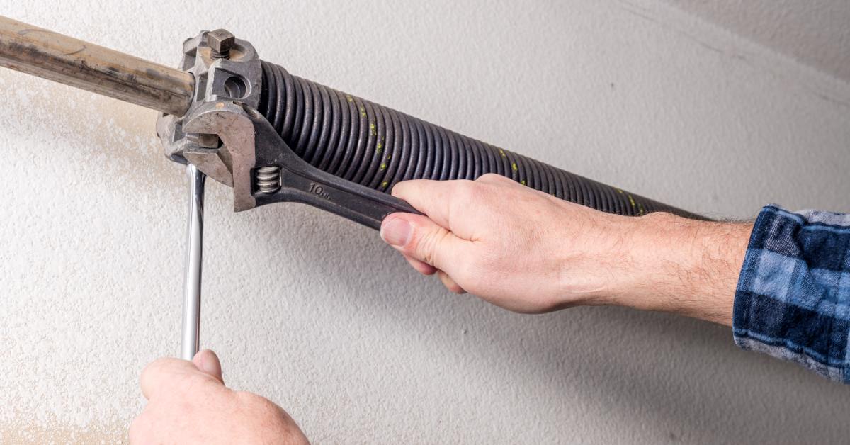 A hand holds a wrench against a coiled garage door spring attached to a metal track on a textured wall.
