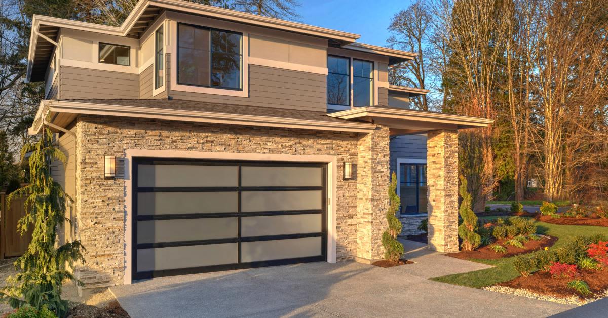 A modern glass garage door with black frames complements a two-story home with tan siding and stone brick accents.