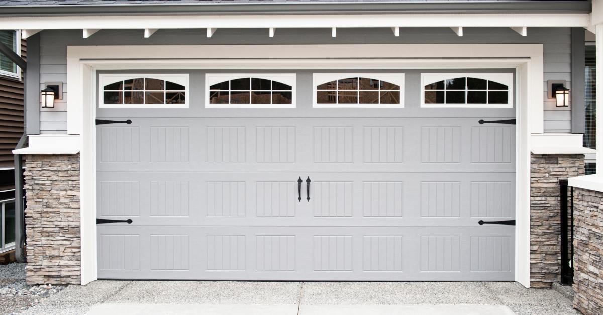 A white carriage-style garage door features arched windows and decorative hardware against a home with stone accents.