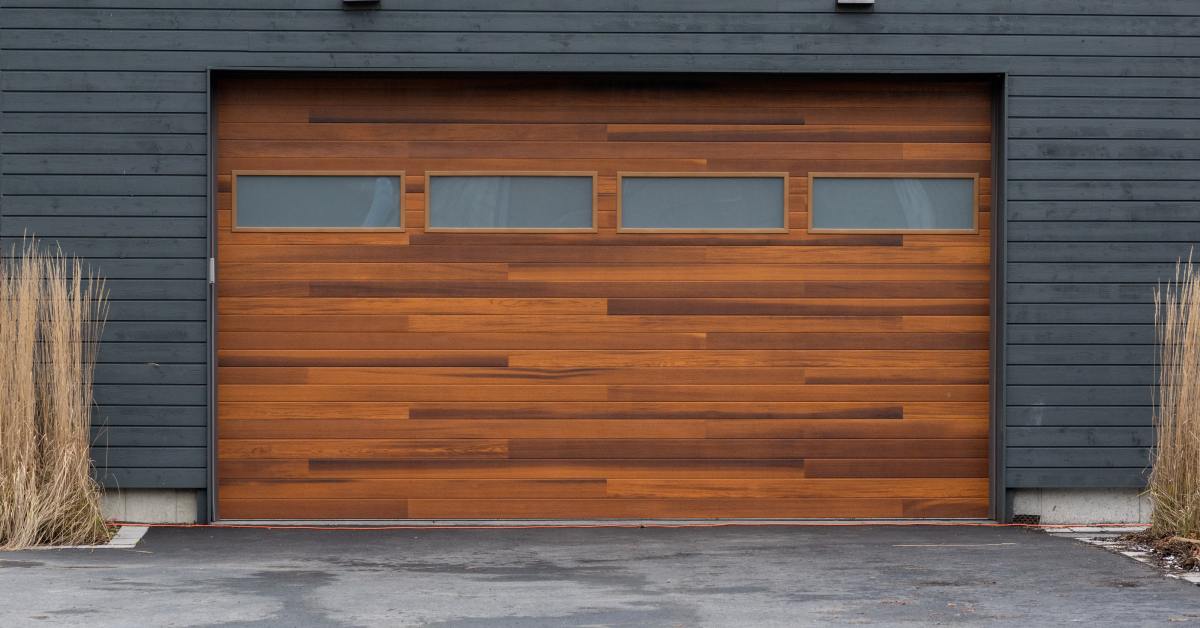 A modern brown wooden garage door with four frosted glass windows sits against a dark gray home exterior.