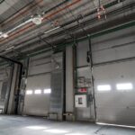 Large sectional steel garage doors lines a clean warehouse, with overhead tracks, pipes, and ceiling lights visible above.