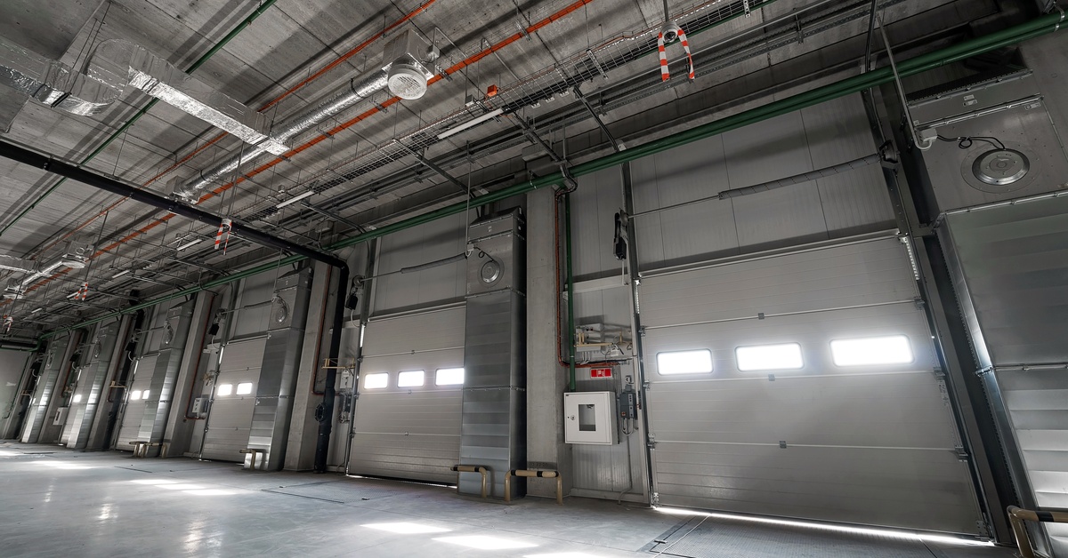Large sectional steel garage doors lines a clean warehouse, with overhead tracks, pipes, and ceiling lights visible above.