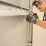 A technician in red overalls and safety glasses adjusts a garage door hinge while inspecting the panel alignment.