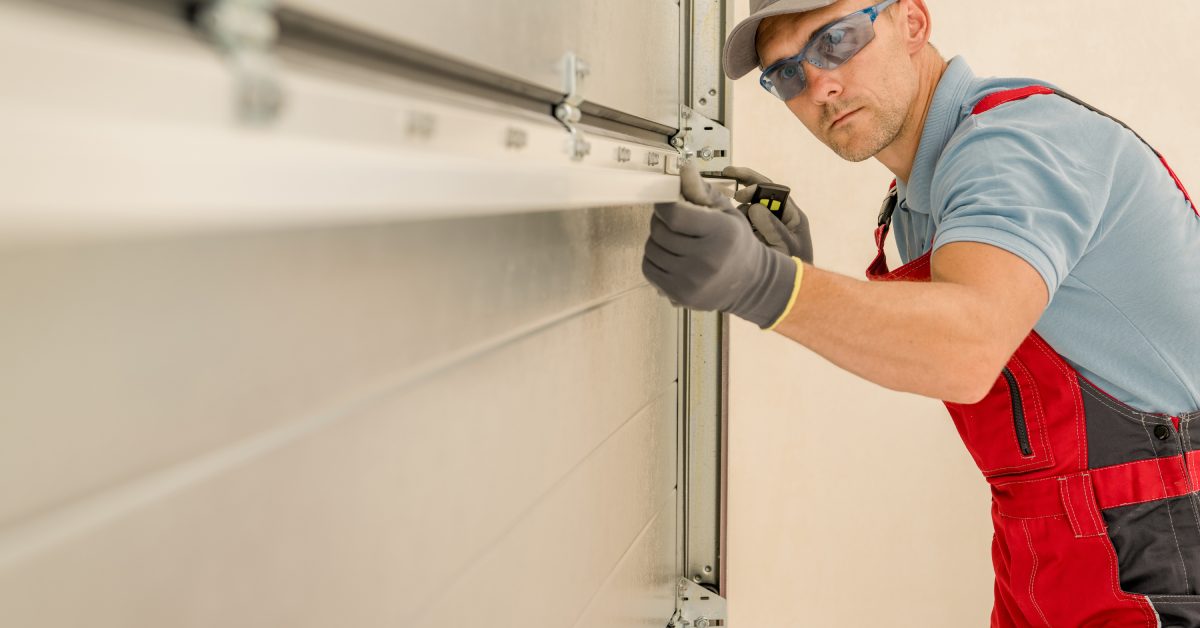 A technician in red overalls and safety glasses adjusts a garage door hinge while inspecting the panel alignment.