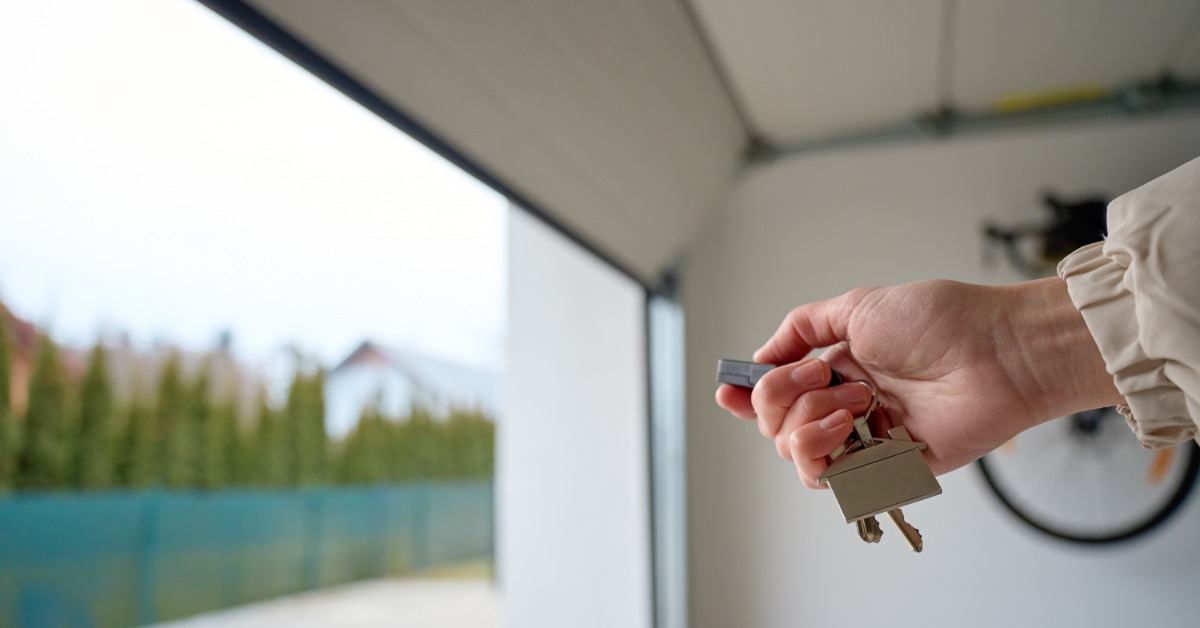 A hand holds a garage door remote and keys while a garage door opens to a driveway with a fence and trees outside