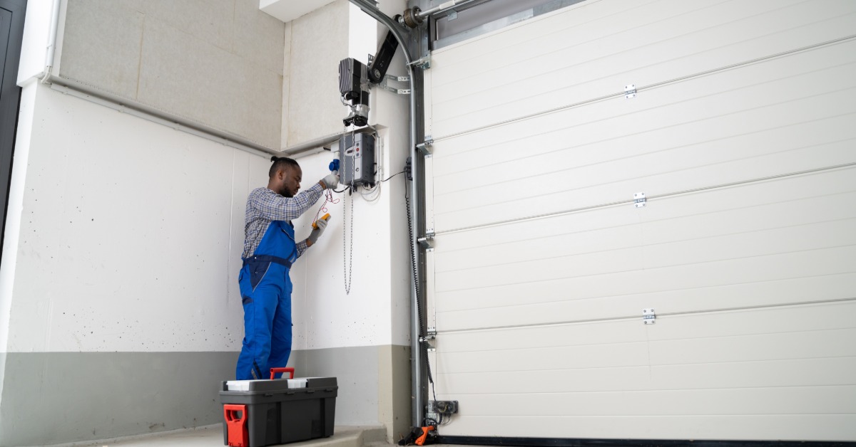 A technician in blue overalls stands on a step and repairs a garage door opener mounted above a closed white door.