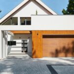 A modern white home features a large wood garage door, clean lines, and a paved driveway framed by green trees.