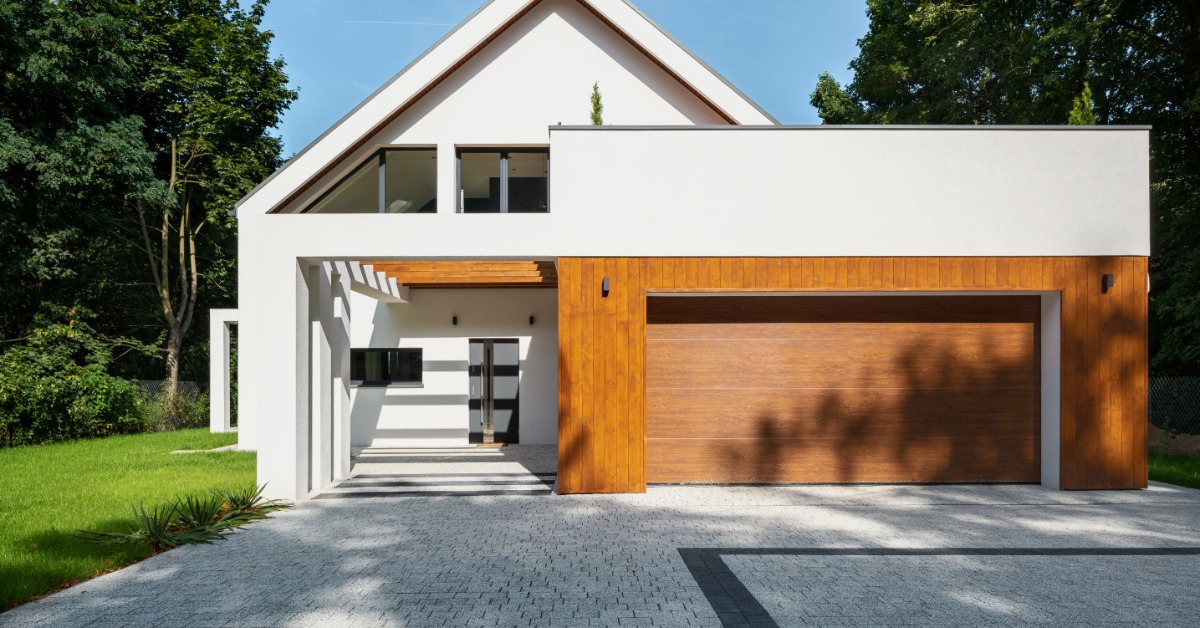 A modern white home features a large wood garage door, clean lines, and a paved driveway framed by green trees.