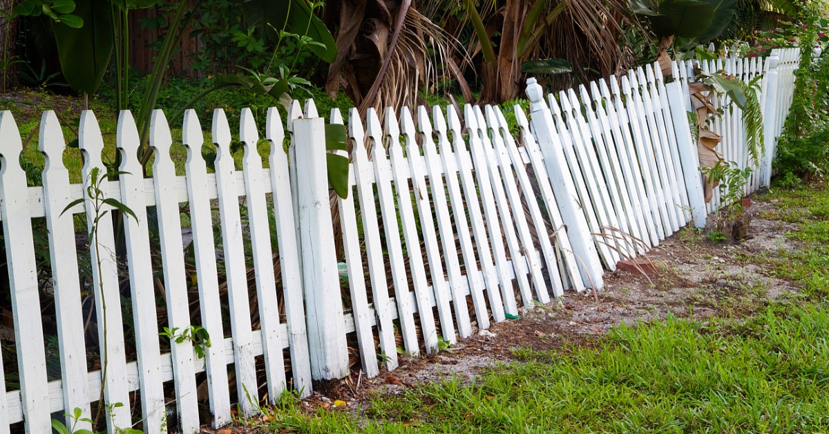 A weathered wooden fence with uneven boards stands in a yard with a tiled roof house and tree branches in the background.